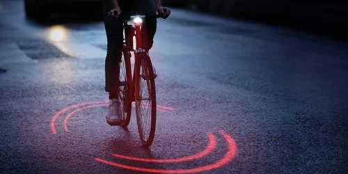 Bicicleta com luz frontal branca e luz traseira projetando dois semicírculos vermelhos no asfalto escuro. Luz e imãs genial.