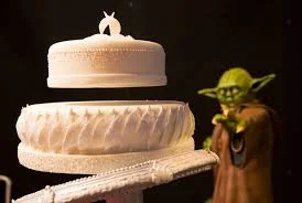 Bolo do Mestre Yoda levita pela Força: um bolo branco de dois andares flutuando, com a figura do Mestre Yoda estendendo a mão.