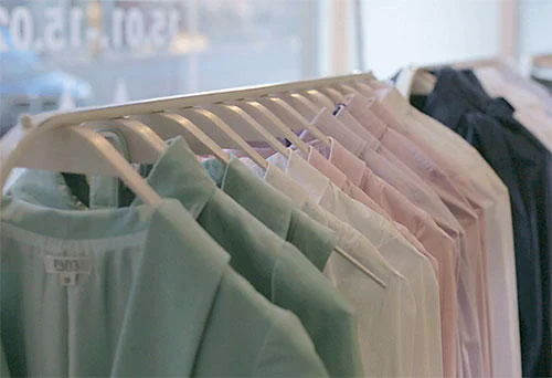 Cabides brancos com camisas coloridas (verde, rosa, branco, azul) alinhadas em arara de loja.