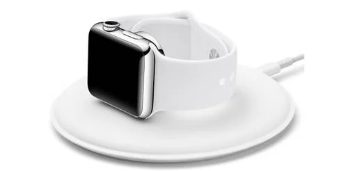 Dock magnética para Apple Watch. Relógio com caixa prateada e pulseira branca apoiado sobre base de carregamento redonda branca.