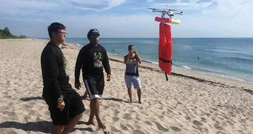 Drone carrega boia de resgate vermelha sobre praia. Três homens observam o drone que ajuda a salvar vítimas de afogamento.