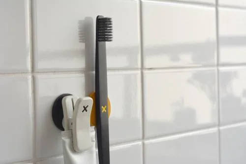 Escova de dente preta com cerdas escuras, presa por ímã redondo amarelo em parede de azulejo branco. Tubo de pasta em clipe.