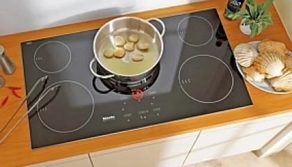 Fogão cooktop por indução magnética Miele preto com 5 zonas, panela com vieiras, pinças e conchas em bancada de madeira.