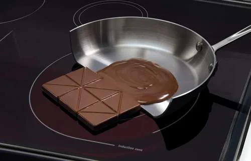 Fogão cooktop de indução magnética preto com panela e chocolate derretido, e um bloco de chocolate sólido ao lado.