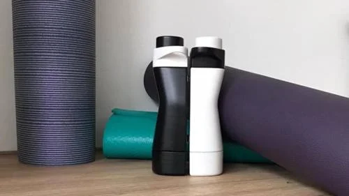 Garrafa de água magnética 2: Duas garrafas, preta e branca, formato ergonômico, lado a lado, entre tapetes de yoga.