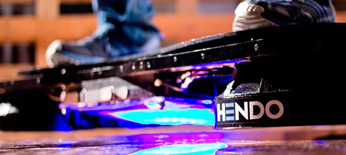 Hoverboard Hendo, o skate voador preto, com luz azul brilhante sob os pés de uma pessoa. Logotipo HENDO visível.