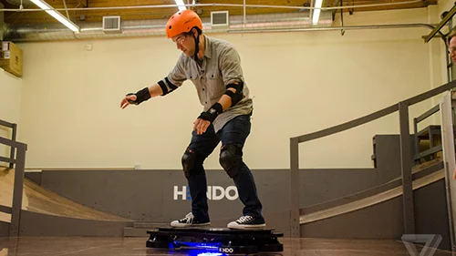 Hoverboard Hendo, o skate voador. Homem de capacete laranja e proteções equilibra-se em Hendo preto flutuante com luz azul em pista coberta.