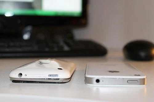 iPhones brancos de modelos diferentes, um com design curvo e outro plano, lado a lado. Detalhe das bordas superiores.