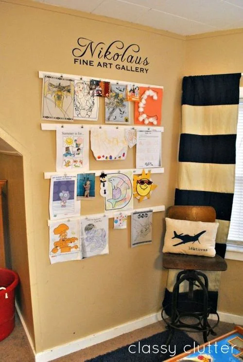 Imãs magnéticos de parede, expondo obras de arte infantis coloridas em varais brancos com o letreiro Nikolaus FINE ART GALLERY.