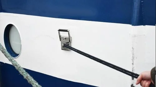 Imã de fixação para navios de cruzeiro: dispositivo metálico cinza com alça, no casco branco e azul. Uma mão conecta um cabo preto a ele.