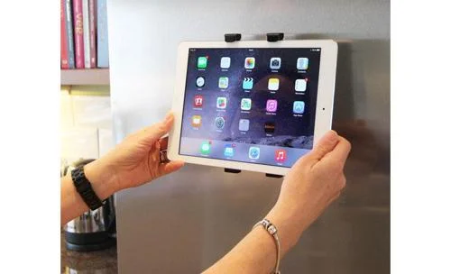 iPad Pro branco, fixado na geladeira de inox por ímãs pretos. Mãos seguram o tablet, exibindo apps na tela.