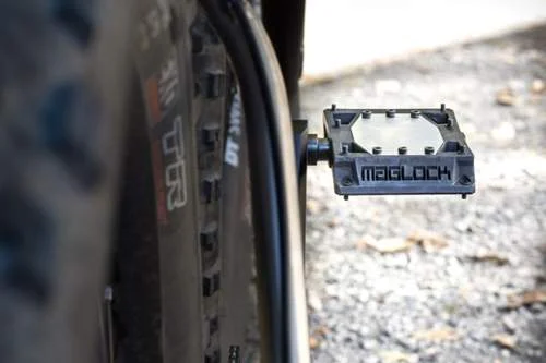 Pedal magnético MAGLOCK preto da Kickstarter para bike. Possui centro claro, pinos e está fixado na bicicleta em chão de cascalho.