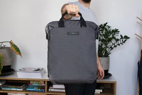 Mochila cinza-escura, vertical, com alças e zíper superior, segurada por pessoa. Design inclui fecho magnético.