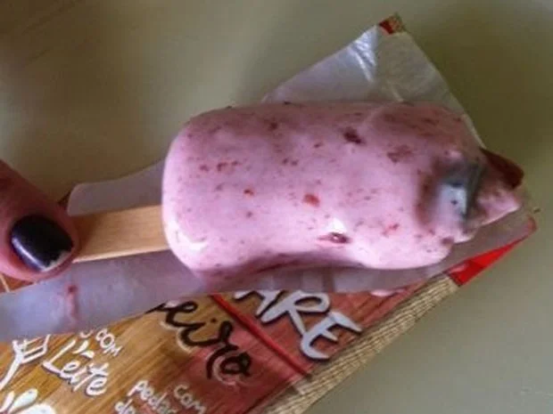 Picolé rosa da Kibon com parafuso metálico visível no interior. Embalagem parcialmente aberta e palito de madeira.