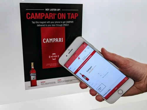 Cartaz Campari on Tap com ímã NFC. Mão usando smartphone para pedido de bebidas Campari em app de entrega.