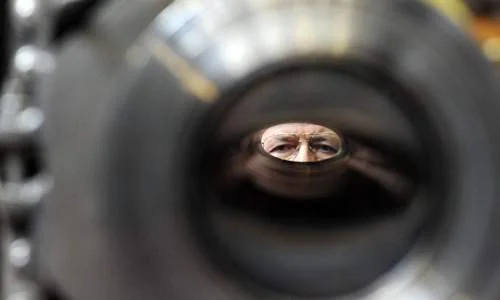 Olhos de um pesquisador, com olhar focado, vistos através de um componente cilíndrico metálico escuro.