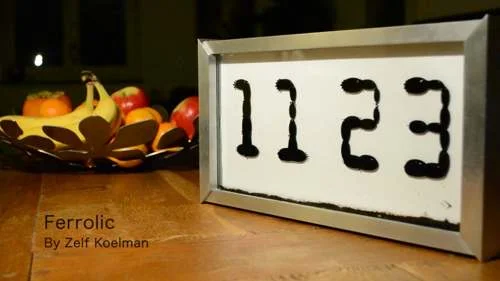 Relógio de ferrofluido digital exibindo 11:23 com fluido magnético preto em moldura prateada, sobre mesa de madeira e fruteira ao fundo.