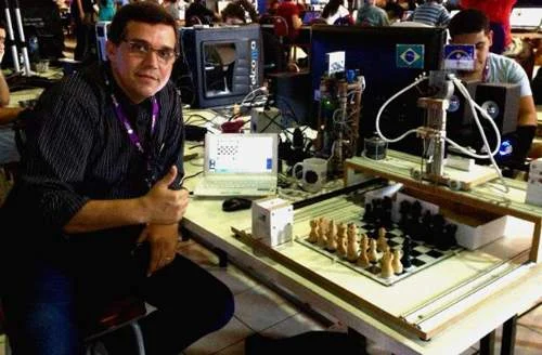 Robô de xadrez com eletroímã em construção, com braço robótico sobre o tabuleiro de peças e homem sorridente.