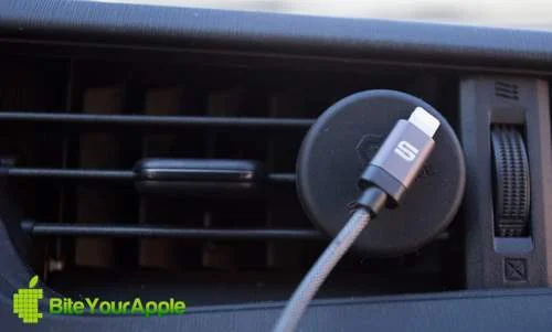 Suporte magnético para smartphones redondo, preto, fixado na saída de ar do carro, com cabo Lightning cinza na frente.