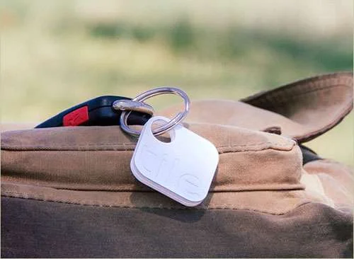 Tile branco, o localizador de objetos perdidos, em chaveiro com controle remoto de carro, sobre tecido marrom.